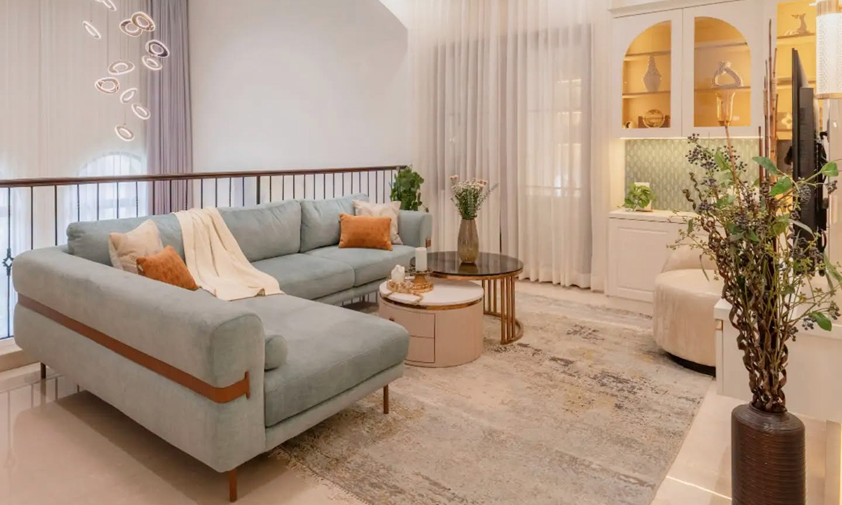 A modern living room featuring a light sage green L-shaped sectional sofa with orange accent pillows, a marble-top nesting coffee table, and soft beige curtains.